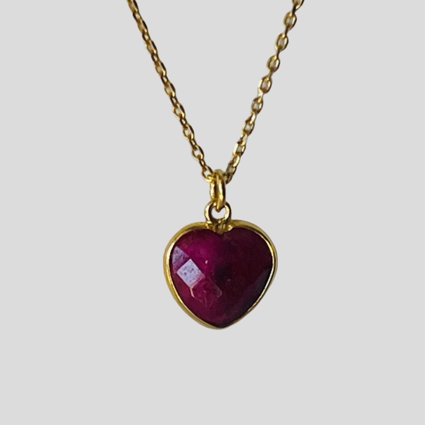 Gold necklace with a heart-shaped red gemstone pendant on a light gray background