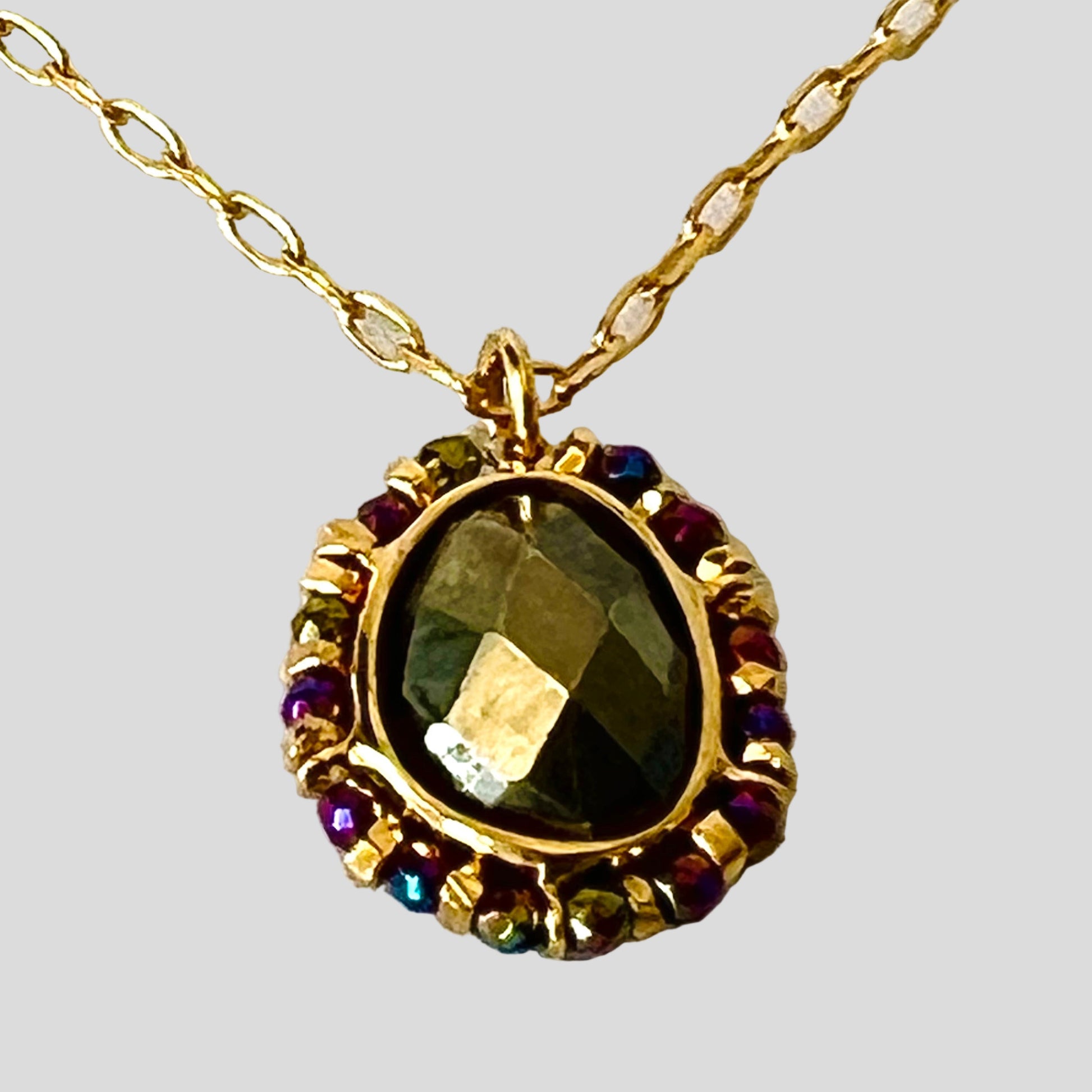 Gold necklace with a pendant featuring a green gemstone and smaller colored stones on a white fabric background.