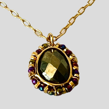 Gold necklace with a pendant featuring a green gemstone and smaller colored stones on a white fabric background.